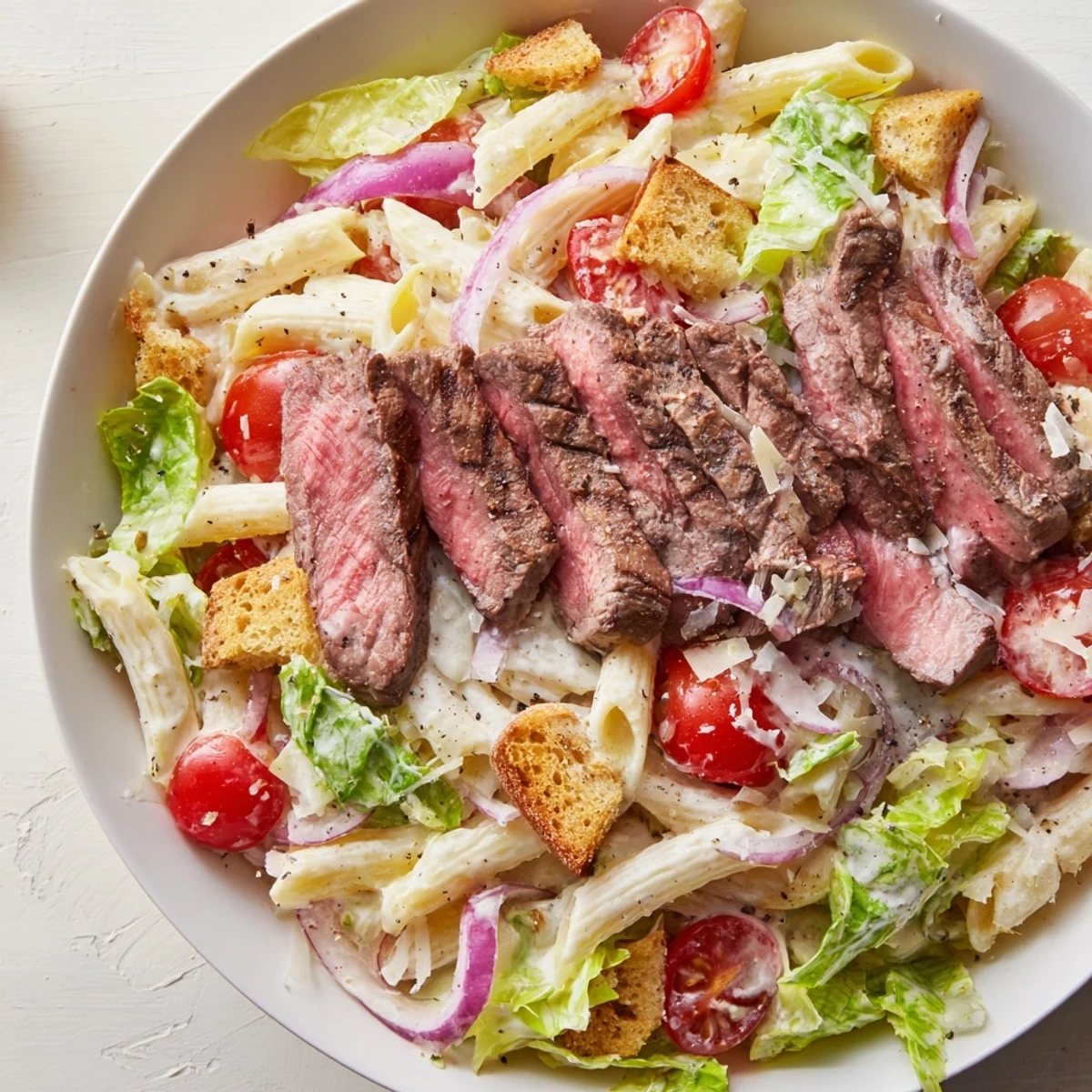 A close-up of Flavorful Steak Caesar Pasta Salad shows juicy grilled steak slices atop crisp romaine and penne.