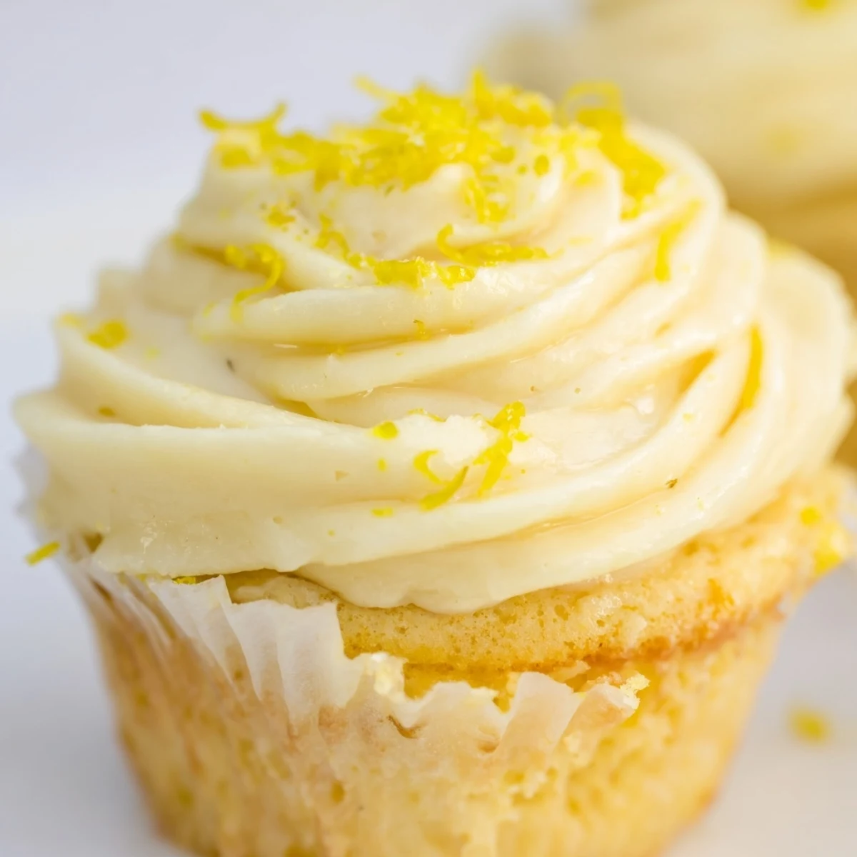 A close-up of a Limoncello cupcake reveals fluffy crumbs, lemon zest garnish, and a pastel yellow glaze drizzle.